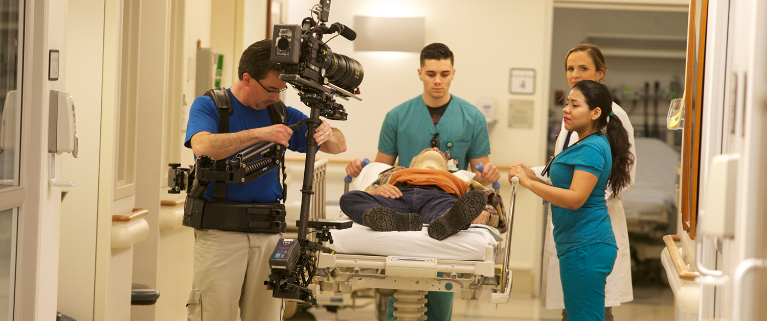 A cameraman films a man laying on a hospital bed.